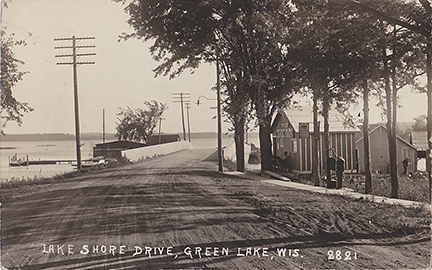 10452-Lake Shore Drive, Green Lake Wisconsin. This card is a view looking south. The Captain Pierce's Boat house is to the right. Looks like Captain Pierce is relaxing by the front door. Norton's Marina can be seen just past the bridge on the lake side. - Postmarked 1921